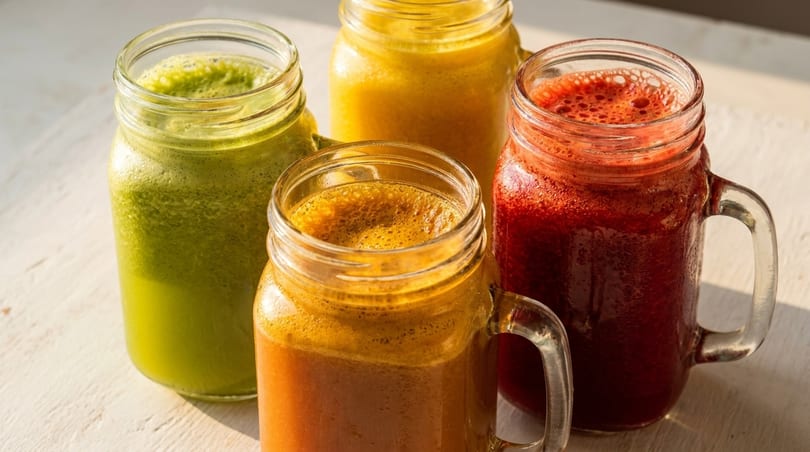 Four mason jars filled with colorful, foamy fresh juices in green, yellow, orange, and red.