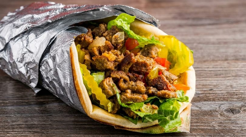 A gyro sandwich, wrapped in foil, filled with meat, lettuce, tomatoes, and pickled peppers.