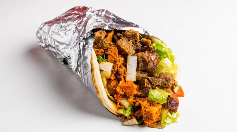 A foil-wrapped gyro, filled with meat, lettuce, onions, and tomatoes, on a white background.